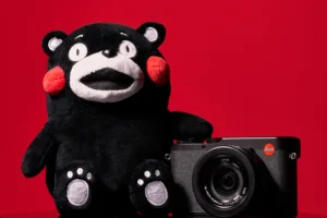 Black Kumamon plush beside a black Leica camera against a solid red background.