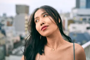 Portrait of a woman in red lipstick and a gold necklace against a Tokyo skyline at dusk.