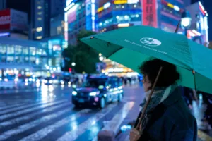 Person with an umbrella at a busy Shibuya crossing at night, city lights and a taxi behind.