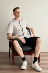 Full-length studio portrait of a man seated in a black leather and wood armchair, wearing a grey short-sleeve shirt, black shorts, and white sneakers, photographed at JG Studio in Tokyo.