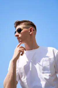 Low-angle outdoor portrait of a man wearing gold-framed sunglasses and a white pocket t-shirt against a clear blue sky, captured during an editorial session in Tokyo by JG.