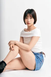 Studio portrait photography in Tokyo of a woman with a short bob seated against a white backdrop, wearing a white off-shoulder top and denim shorts, soft studio lighting.