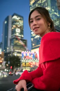 Photographie de portrait lors d'une visite nocturne à Shibuya, modèle en pull rouge devant les panneaux publicitaires illuminés du carrefour de Shibuya et la skyline en arrière-plan.