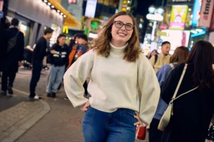 Femme souriante en col roulé blanc posant dans une rue animée de Shibuya la nuit, entourée d'enseignes au néon, lors de la balade photo de JG à Shibuya.